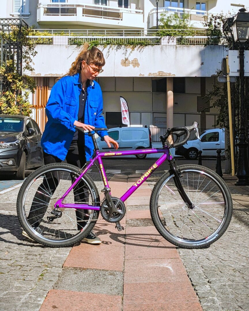 Photo d'une mécanicienne de La Fabrique des Cyclistes présentant un gravel d'occasion dans la rue devant la boutique.