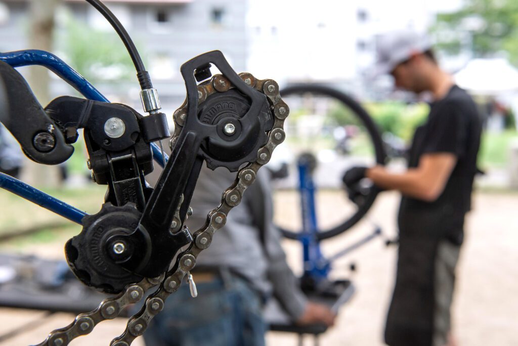 Photo d'un dérailleur de vélo en gros plan. Un mécanicien de La Fabrique des Cyclistes travaille sur un autre vélo en second plan.