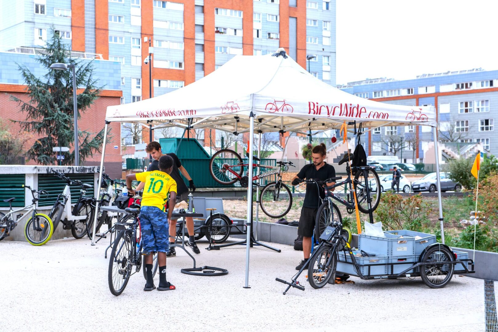 Photo de deux mécaniciens de La Fabrique des Cyclistes travaillant sur des vélos sous une tente d'atelier mobile au milieu de la ville. Un enfant avec son vélo leur parle.