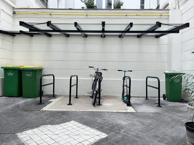 Photo de 5 arceaux de stationnement vélo abrité par un auvent en verre dans la cour d'un immeuble.