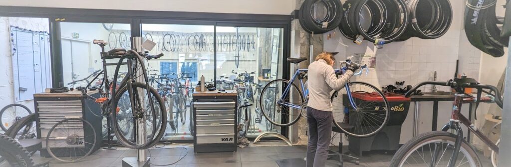 Photo de l'atelier de La Fabrique des Cyclistes où une mécanicienne travaille sur un vélo et où des rangées de vélos et de roues est visible en fond à travers une baie vitrée.