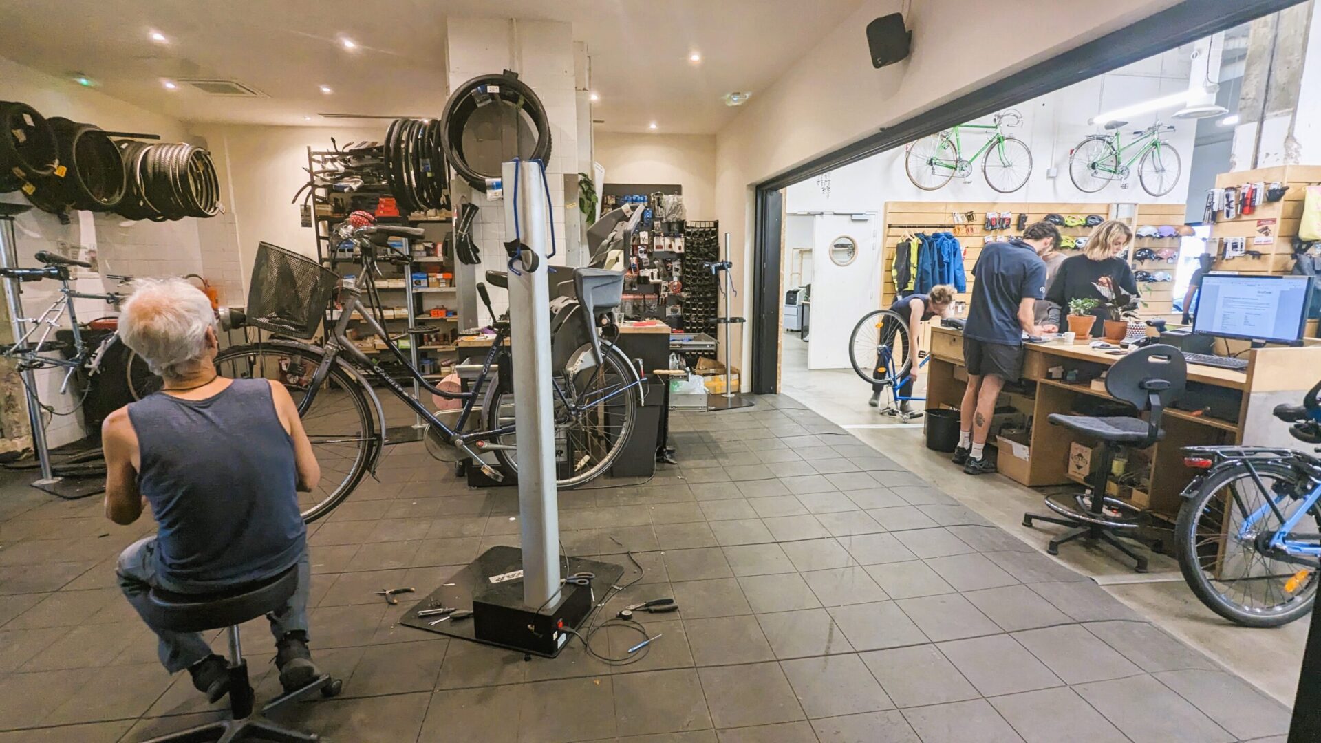 Photo de l'atelier-boutique de La Fabrique des Cyclistes montrant en grand angle la partie atelier à gauche avec un mécanicien affairé, et la partie boutique à droite où un mécanicien accueille une cliente.