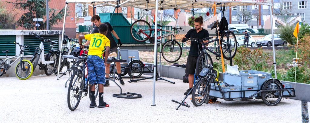 Photo de deux mécaniciens de La Fabrique des Cyclistes travaillant sur des vélos sous une tente d'atelier mobile au milieu de la ville. Un enfant avec son vélo leur parle.