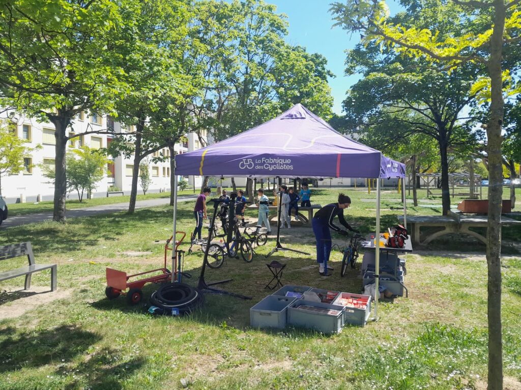 Photo d'un atelier mobile installé sous un barnum violet dans un parc aux pieds d'un immeuble.