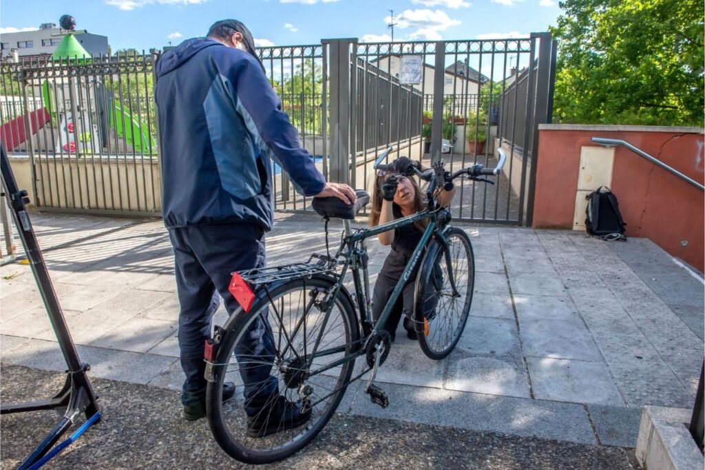 Photo d'une mécanicienne réglant les freins d'un vélo lors d'un atelier mobile devant un portail.
